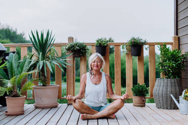 Senior woman sitting outdoors on a terrace in summer, doing yoga exercise. - HPIF11986