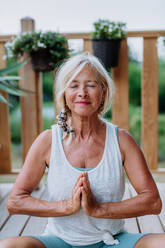 Ältere Frau, die im Sommer auf einer Terrasse sitzt und Yogaübungen macht. - HPIF11990