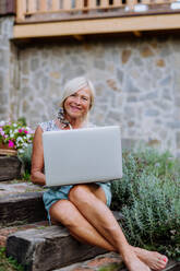 Eine ältere Frau benutzt einen Laptop und bearbeitet Bestellungen für ihre selbst angebauten Bio-Blumen und Gemüse im Garten - HPIF12003