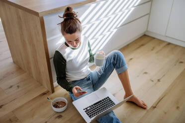 Junge Frau sitzt mit Laptop und einer Tasse Kaffee in ihrer Küche. - HPIF12043