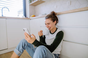 Junge Frau sitzt mit Smartphone und einer Tasse Kaffee in ihrer Küche. - HPIF12046