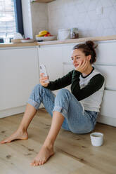 Junge Frau sitzt mit Smartphone und einer Tasse Kaffee in ihrer Küche. - HPIF12047