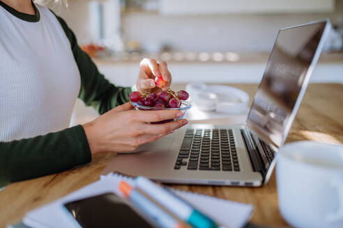 Seitenansicht einer Frau, die eine Schale mit Weintrauben über einem Schreibtisch mit Computer, Tagebuch und Smartphone hält, Konzept der Work-Life-Balance. - HPIF12071