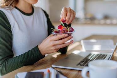 Side view of woman holding bowl with grapes above a desk with computer, diary and smartphone. Work-life balance concept. - HPIF12073