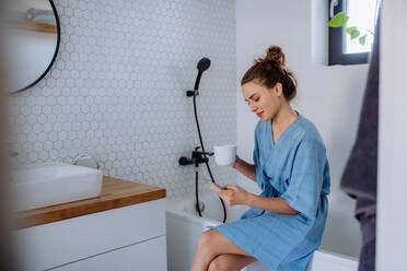 Young woman in bathroom with phone and cup of coffee, morning routine and relaxation concept. - HPIF12129