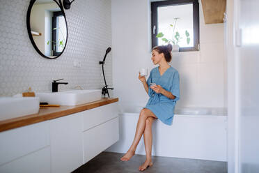 Young woman in bathroom with phone and cup of coffee, morning routine and relaxation concept. - HPIF12130