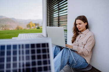 Junge Frau sitzt auf der Terrasse und lädt ihr Tablet mit dem Solarpanel auf. - HPIF12154