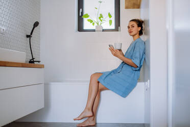 Young woman in bathroom with phone and cup of coffee, morning routine and relaxation concept. - HPIF12163