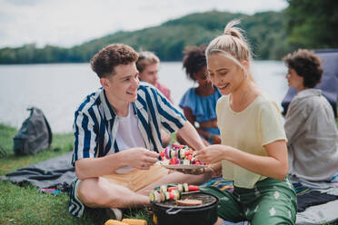 Eine Gruppe multirassischer junger Freunde zeltet auf einem Campingplatz in der Nähe eines Sees und grillt, indem sie Spieße auf den Grill legen. - HPIF12226