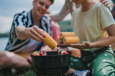 A close-up of young friends putting corn on grill and having barbecue when camping in campground. - HPIF12228