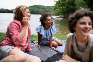 A diverse group of friends having fun together at park lauging and sitting on blanket near lake, Friendship and lifestyle concepts - HPIF12238