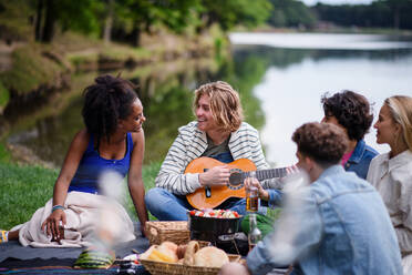 Eine Gruppe junger Freunde vergnügt sich bei einem Picknick in der Nähe eines Sees, sitzt auf einer Decke, isst und spielt Gitarre. - HPIF12243