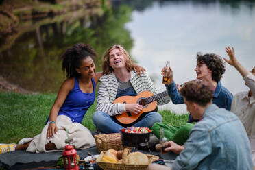 A group of young friends having fun on picnic near a lake, sitting on blanket eating and playing guitar. - HPIF12244