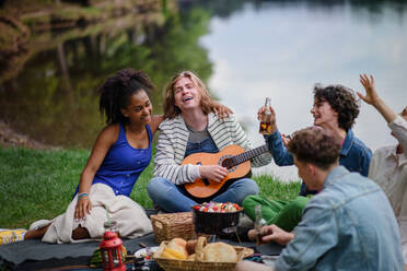 Eine Gruppe junger Freunde vergnügt sich bei einem Picknick in der Nähe eines Sees, sitzt auf einer Decke, isst und spielt Gitarre. - HPIF12244