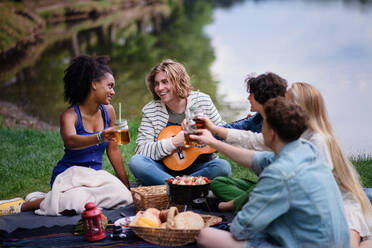 Eine Gruppe junger Freunde vergnügt sich bei einem Picknick in der Nähe eines Sees, sitzt auf einer Decke und stößt mit Getränken an. - HPIF12245