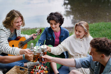 Eine Gruppe junger Freunde vergnügt sich bei einem Picknick in der Nähe eines Sees, sitzt auf einer Decke und stößt mit Getränken an. - HPIF12247