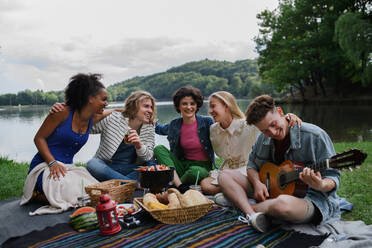 Eine Gruppe junger Freunde vergnügt sich bei einem Picknick in der Nähe eines Sees, sitzt auf einer Decke, isst und spielt Gitarre. - HPIF12250