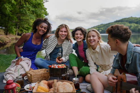 Eine Gruppe multirassischer junger Freunde zeltet in der Nähe eines Sees und grillt gemeinsam, während sie in die Kamera schauen. - HPIF12251