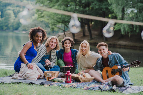 Eine Gruppe junger Freunde vergnügt sich bei einem Picknick in der Nähe eines Sees, sitzt auf einer Decke, isst und spielt Gitarre. - HPIF12252