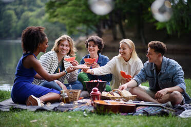 Eine Gruppe junger Freunde vergnügt sich bei einem Picknick in der Nähe eines Sees, sitzt auf einer Decke und isst Wassermelone. - HPIF12255