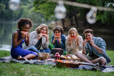 Eine Gruppe junger Freunde vergnügt sich bei einem Picknick in der Nähe eines Sees, sitzt auf einer Decke und isst Wassermelone. - HPIF12256