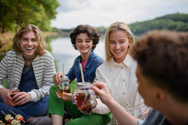 Eine Gruppe junger Freunde vergnügt sich bei einem Picknick in der Nähe eines Sees, sitzt auf einer Decke und stößt mit Getränken an. - HPIF12258