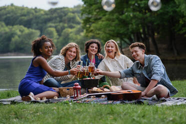 Eine Gruppe junger Freunde vergnügt sich bei einem Picknick in der Nähe eines Sees, sitzt auf einer Decke und stößt mit Getränken an und schaut in die Kamera. - HPIF12261