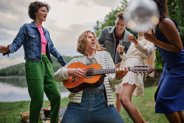 Eine Gruppe junger Freunde vergnügt sich bei einem Picknick an einem See, lacht, tanzt und spielt Gitarre. - HPIF12268
