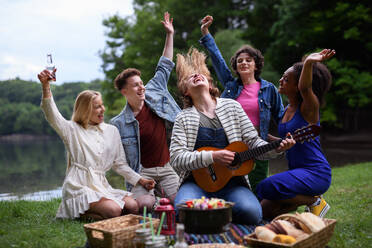 Eine Gruppe junger Freunde vergnügt sich bei einem Picknick in der Nähe eines Sees, sitzt auf einer Decke, isst und spielt Gitarre. - HPIF12270