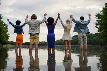 Eine gemischtrassige Gruppe junger Freunde, die sich an den Händen halten und im Sommer in einem See stehen, Rückansicht. - HPIF12281