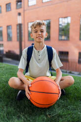 Fröhlicher kaukasischer Junge, der im öffentlichen Stadtpark mit einem Basketball sitzt und in die Kamera schaut. - HPIF12318