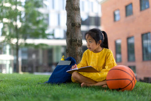 Kleines asiatisches Mädchen sitzt in einem öffentlichen Park und schreibt einige Notizen. Sommerzeit. - HPIF12325