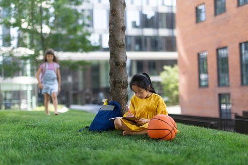 Kleines asiatisches Mädchen sitzt in einem öffentlichen Park und schreibt einige Notizen, ihr Freund steht im Hintergrund. Sommerzeit. - HPIF12327