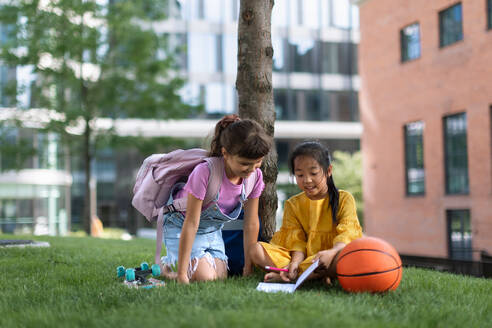 Ein kleines asiatisches Mädchen sitzt mit ihrer Freundin in einem öffentlichen Park und schreibt einige Notizen. Sommerzeit. - HPIF12329