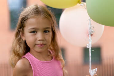 Porträt eines kleinen blonden Mädchens, das mit Luftballons in der Stadt posiert und in die Kamera schaut. - HPIF12341