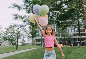 Porträt eines kleinen blonden Mädchens, das mit Luftballons in der Stadt posiert und in die Kamera schaut. - HPIF12343