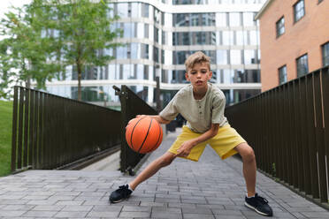 Fröhlicher kaukasischer Junge dribbelt mit einem Basketball im öffentlichen Stadtpark und schaut in die Kamera. - HPIF12346