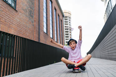 Fröhlicher kaukasischer Junge, der auf einem Skateboard sitzt und den Hügel hinunterfährt und in die Kamera schaut. - HPIF12349