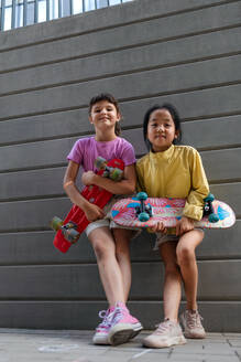 Happy friends posing together in city, standing in front of a concrete wall, holding skateboards and looking at camera. - HPIF12352