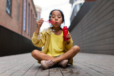 Little girl sitting in a city and blowing bubbles. - HPIF12364