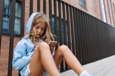 Kleines blondes Mädchen mit Kapuze sitzt auf dem Bürgersteig, hält ein Smartphone und schaut in die Kamera. - HPIF12366