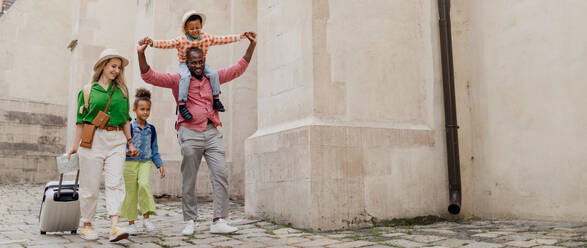 Happy multiracial family travel together with suitcases, walking in old city centre. - HPIF12402