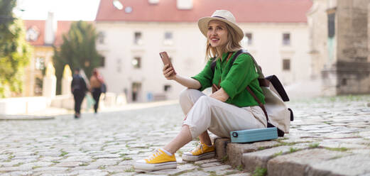 Junge blonde Frau reist allein im alten Stadtzentrum, sitzt und benutzt ihr Smartphone. - HPIF12415