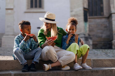 Multirassische Kinder mit Mutter essen eine Wassermelone auf der Straße während der heißen sonnigen Tagen, Sommerurlaub Reisen Konzept. - HPIF12418