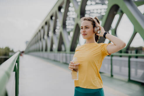Junge Frau trinkt Wasser und hört Musik über Kopfhörer, beim Joggen in der Stadt, gesunder Lebensstil und Sportkonzept. - HPIF12448