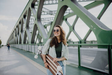 Junge Frau, die mit einem Skateboard auf einer Brücke in der Stadt spazieren geht, Jugendkultur und Pendeln. - HPIF12467