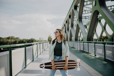 Junge Frau, die mit einem Skateboard auf einer Brücke in der Stadt spazieren geht, Jugendkultur und Pendeln. - HPIF12471