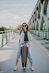 Junge Frau, die mit einem Skateboard auf einer Brücke in der Stadt spazieren geht, Jugendkultur und Pendeln. - HPIF12475