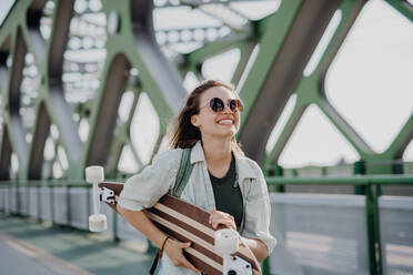 Junge Frau, die mit einem Skateboard auf einer Brücke in der Stadt spazieren geht, Jugendkultur und Pendeln. - HPIF12477