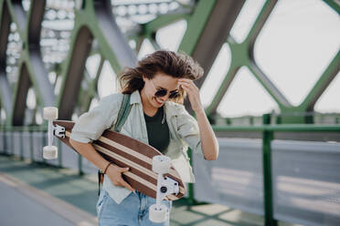 Junge Frau, die mit einem Skateboard auf einer Brücke in der Stadt spazieren geht, Jugendkultur und Pendeln. - HPIF12478